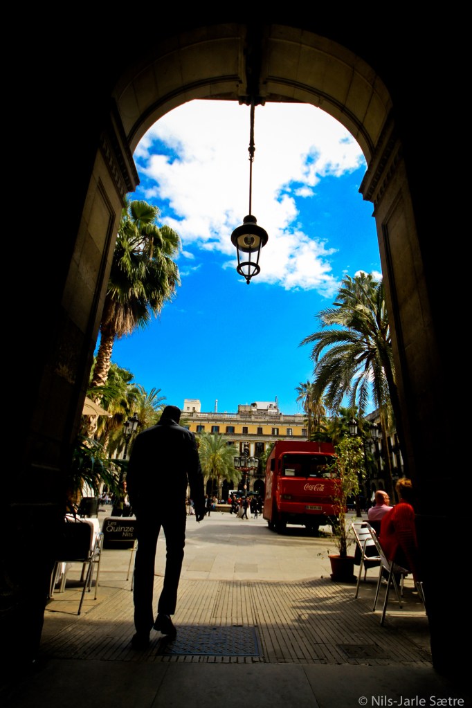 En mann er på vei ut av skyggen og inn i solen på Placa Reial.